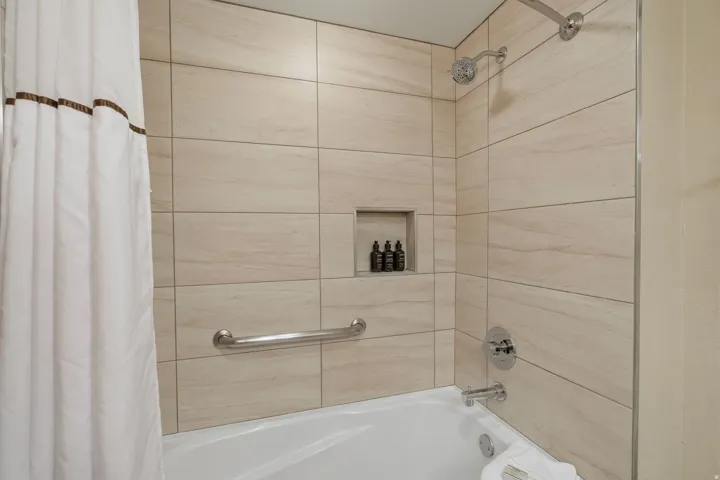 Bathtub with light-toned tiled surround, built-in recessed shelf, chrome shower fixtures, and a grab bar