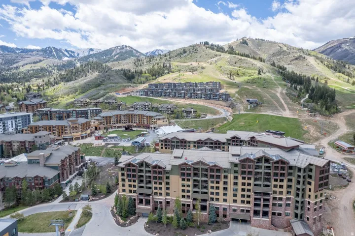 Mountain resort community featuring multi-story buildings with varied architectural styles, a backdrop of snow-capped mountains, and extensive green slopes