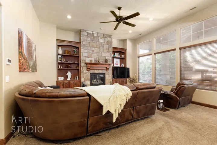 Living area featuring a stone fireplace with a wood mantel, built-in shelving, a ceiling fan, and multiple windows with blinds