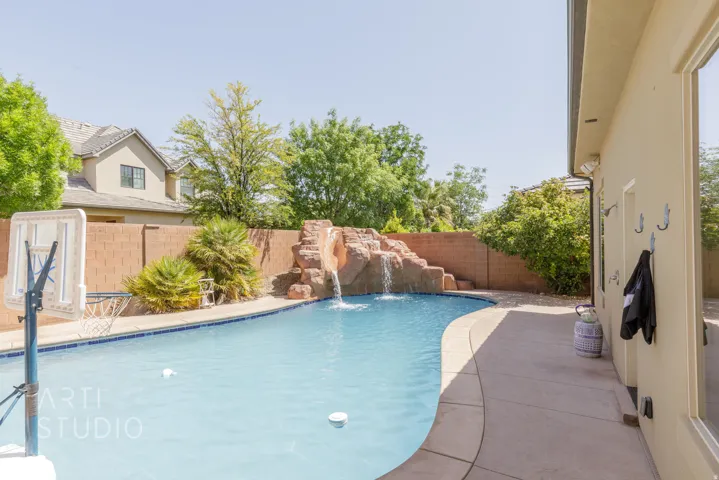 Curved swimming pool featuring a rock waterfall, surrounded by a concrete patio and block wall fencing
