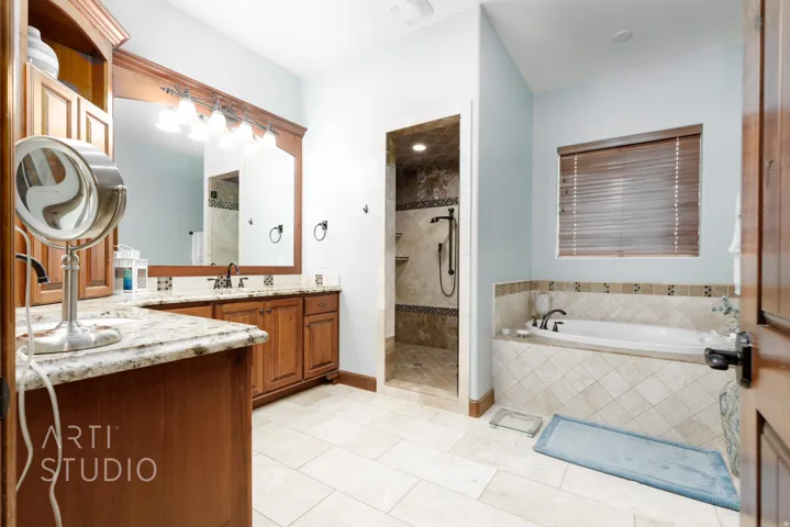 Expansive bathroom featuring a built-in soaking tub with tiled surround, a separate walk-in shower with tiled walls and flooring, and a double vanity with granite-look countertops and wood cabinetry