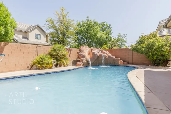 Curved swimming pool featuring a rock waterfall, surrounding concrete patio, mature landscaping, and a privacy wall