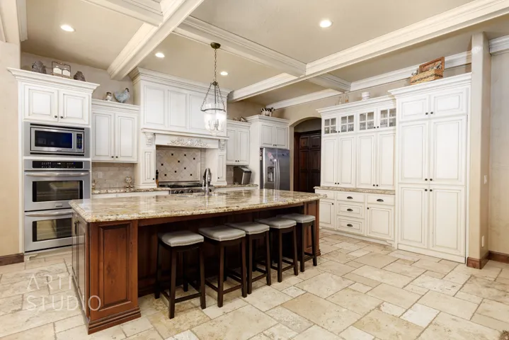 Spacious kitchen featuring an expansive center island with seating, natural stone flooring, white cabinetry with dark hardware, stainless steel built-in appliances, and a coffered ceiling