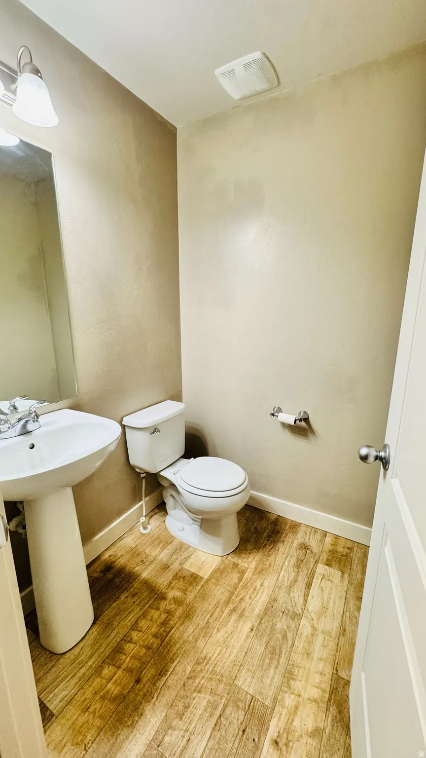 Powder room featuring wood-finish flooring, a white pedestal sink, a toilet, a wall-mounted light fixture, and a ceiling-mounted ventilation fan