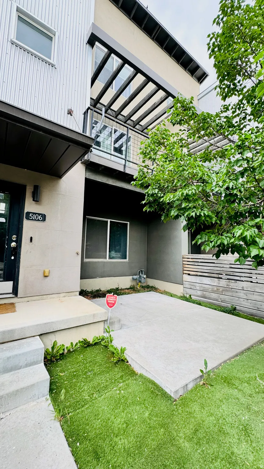 Modern multi-level exterior featuring light-colored stucco, corrugated metal siding, and a second-story balcony with a slatted pergola