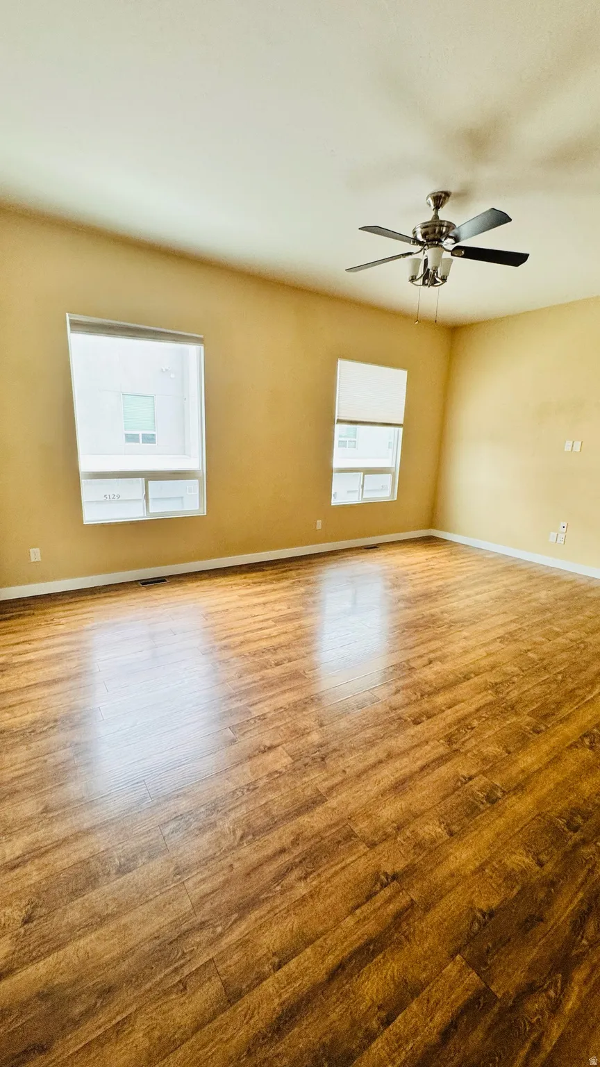 Spacious room featuring wood-finish flooring, light-toned walls, two windows with roller blinds, and a ceiling fan with integrated lighting