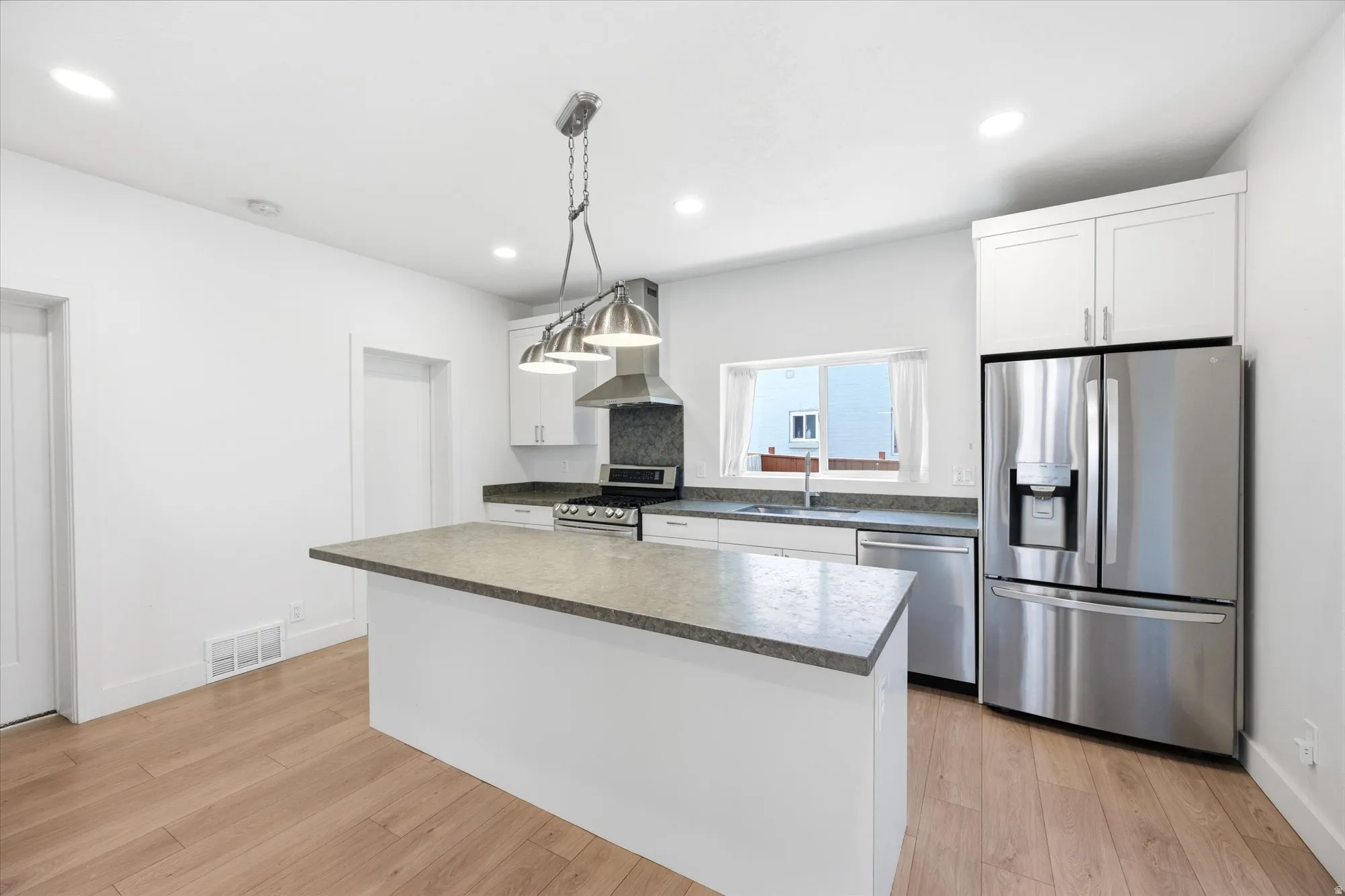 Modern kitchen featuring a central island with a stone-finish countertop, stainless steel appliance suite, white cabinetry, wood-finish flooring, and recessed ceiling lighting
