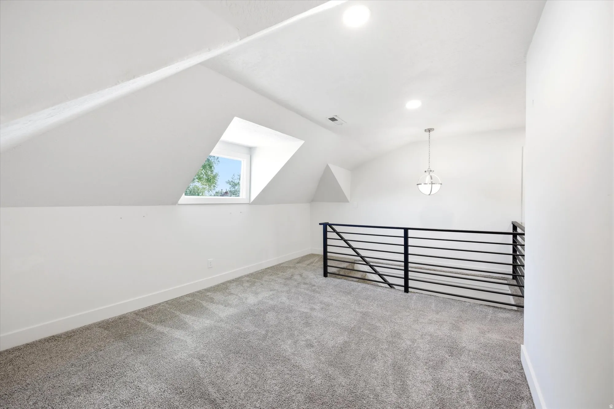 Carpeted loft space featuring a horizontal black metal railing, a prominent dormer window, an overhead pendant light fixture, and recessed lighting