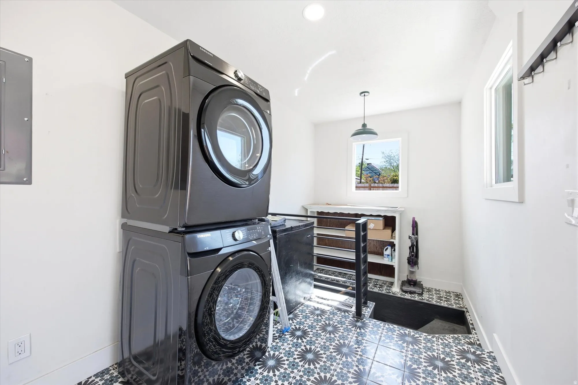 Laundry area featuring a stacked washer and dryer, patterned floor tile, white walls, and a single window with a pendant light fixture