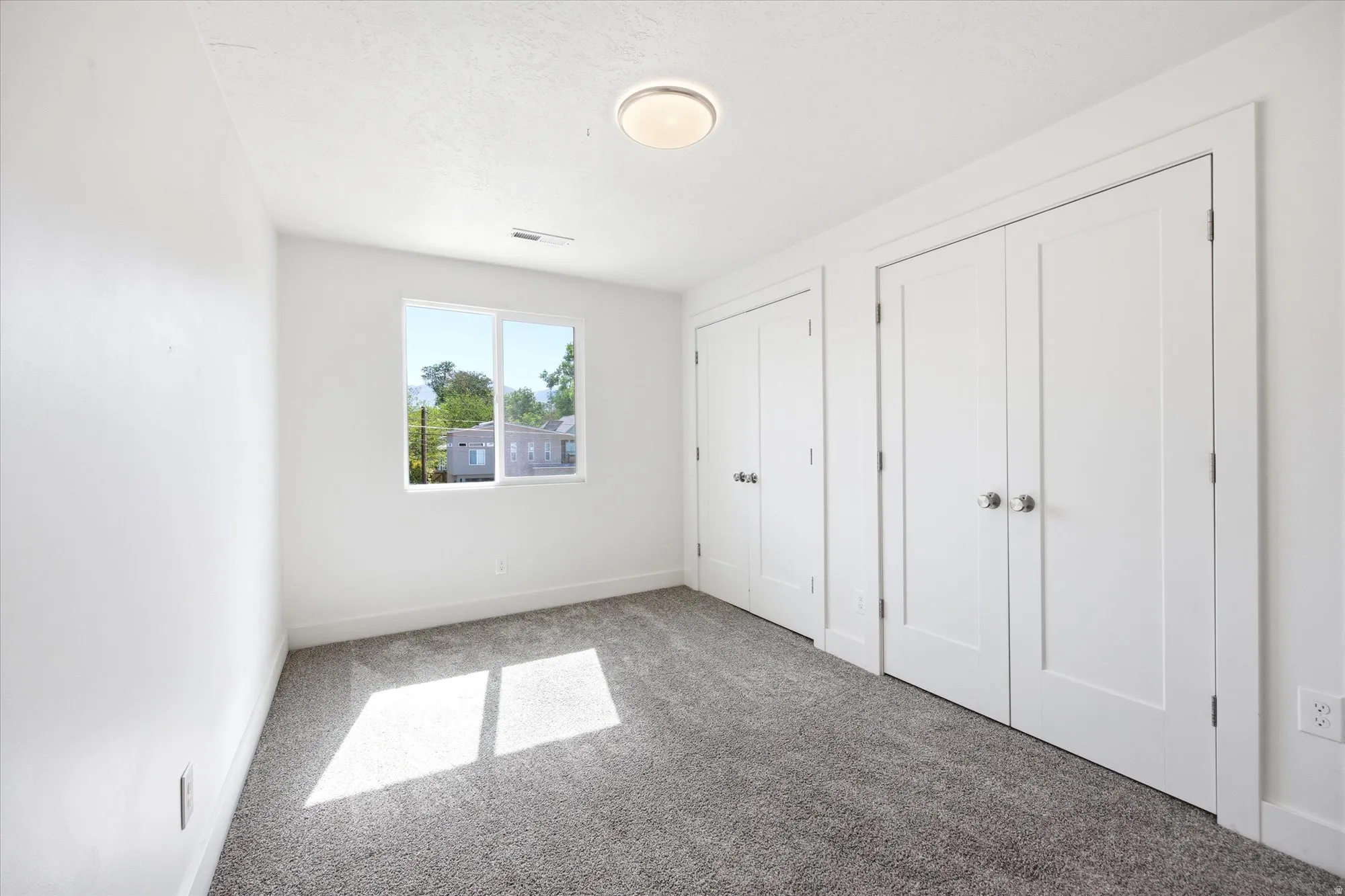 White walls and gray textured carpeting create a neutral palette in this room, featuring two sets of bi-fold closet doors, a double-pane window, and a ceiling-mounted light fixture