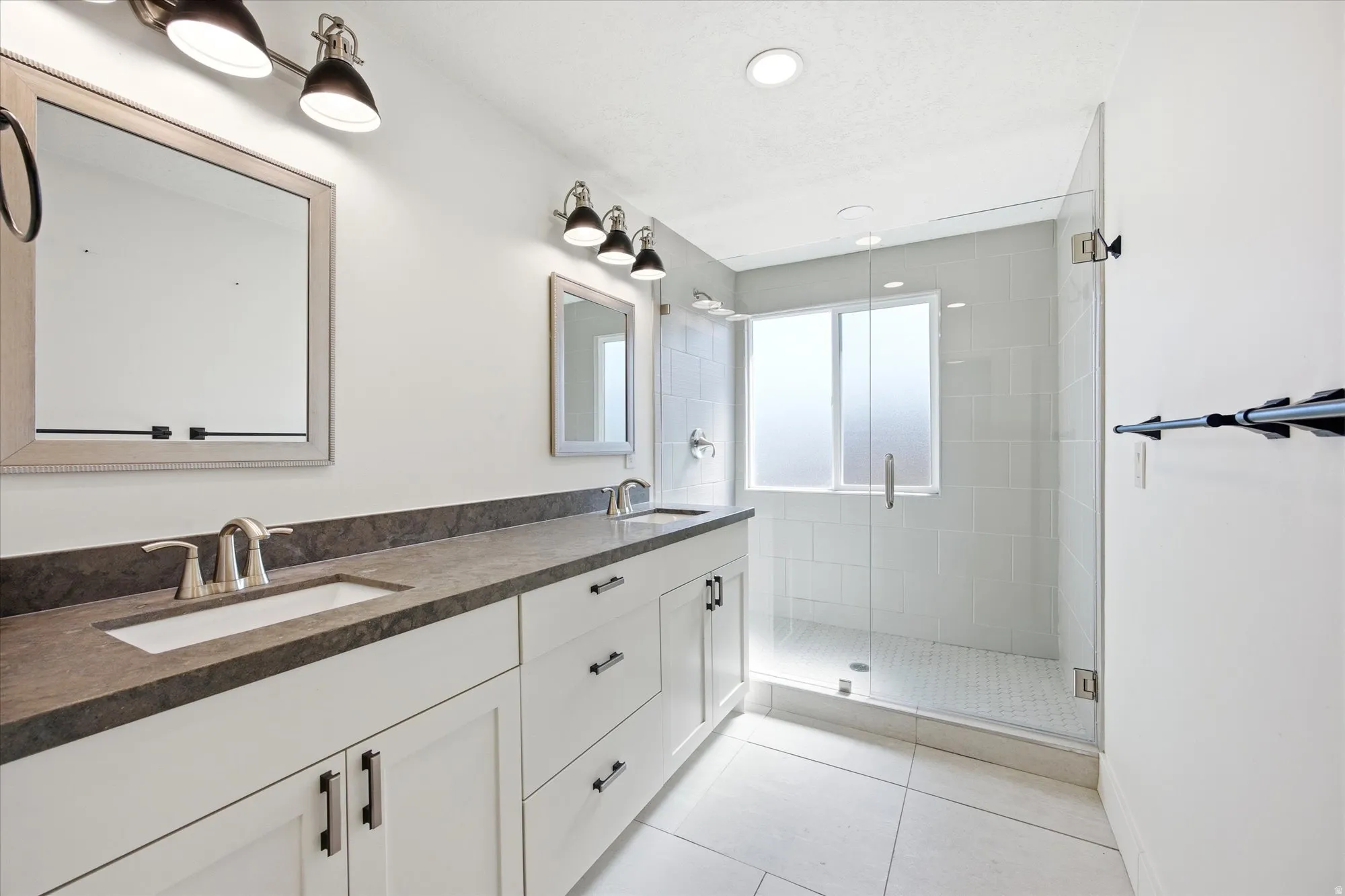 Bathroom featuring a double vanity with dark countertops and integrated sinks, brushed nickel fixtures, white shaker-style cabinetry with dark hardware, and track lighting