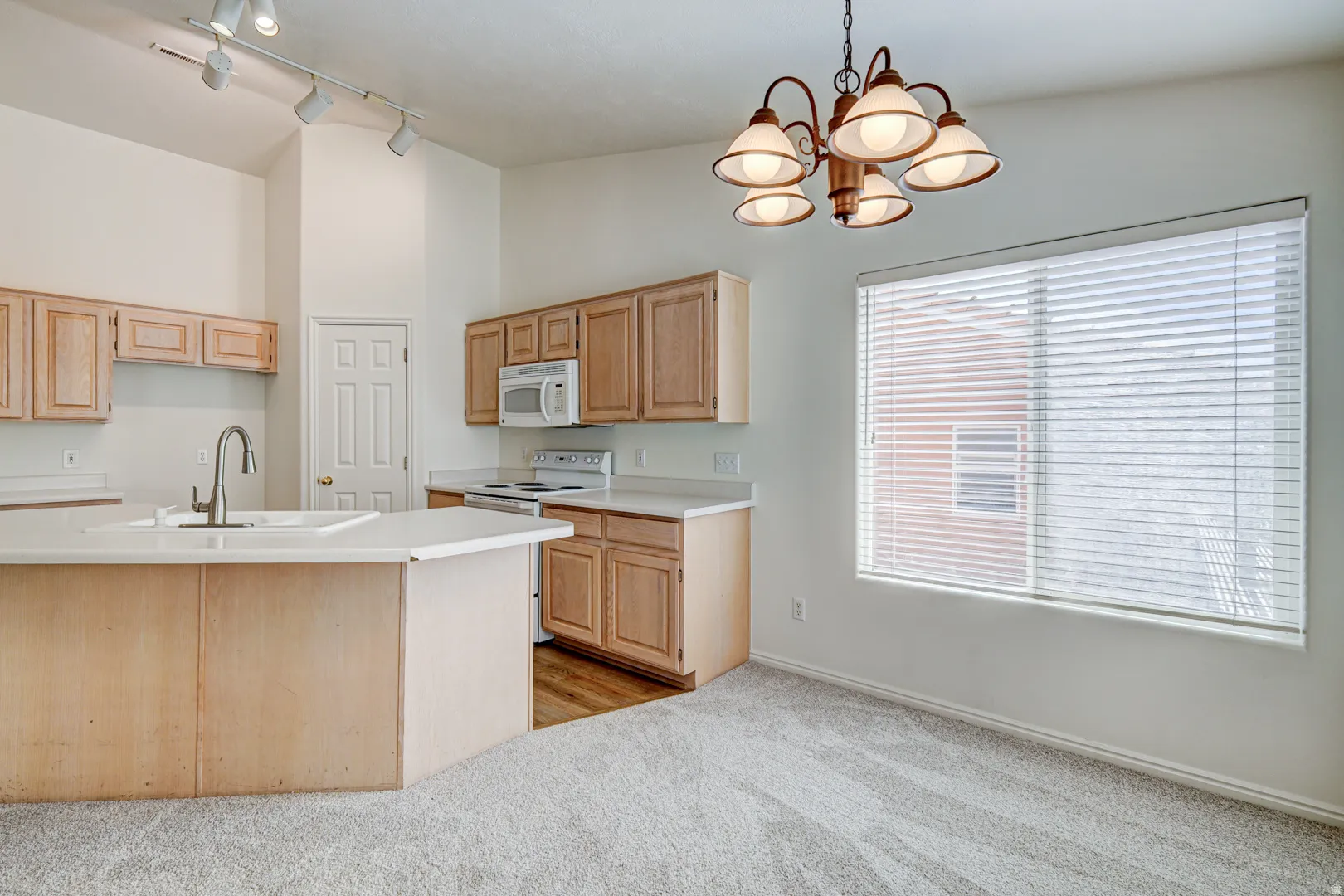 Open-concept kitchen and dining area featuring light wood cabinetry, a central island with an integrated sink, white appliances, and a window with blinds