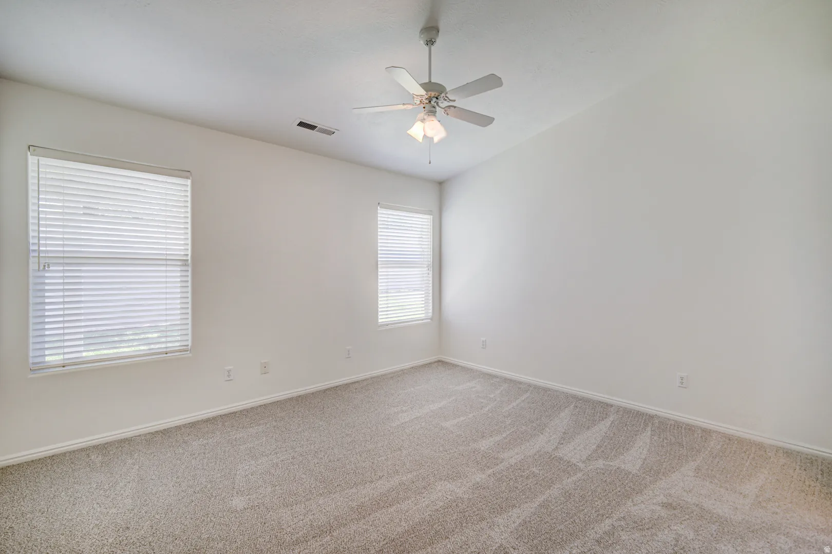 Spacious Primary Suite featuring neutral carpet flooring, white walls, two windows with blinds, a ceiling fan with light fixture, and vaulted ceilings