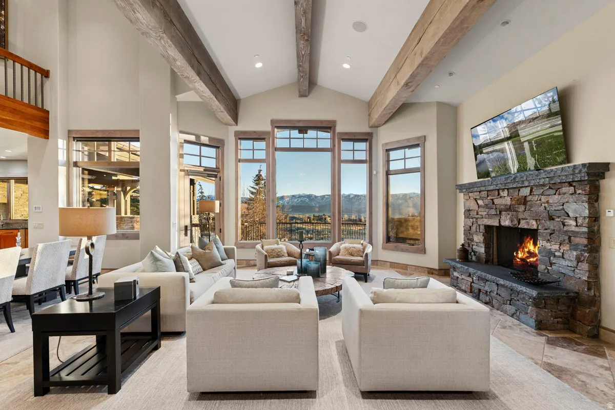 Vaulted ceiling with exposed wood beams, floor-to-ceiling windows, stone fireplace with hearth, and tiled flooring