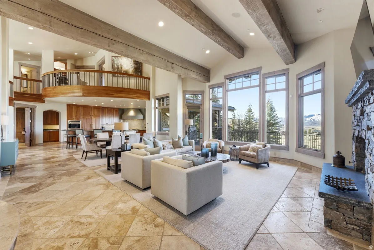 Spacious living area featuring exposed wood beams, a stone fireplace, large picture windows, and patterned tile flooring