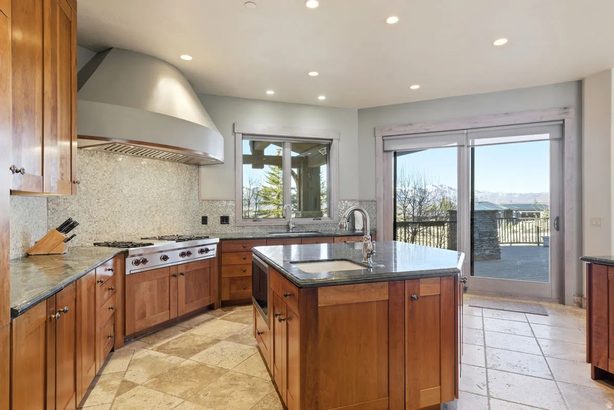 Hardwood cabinetry kitchen featuring a center island with undermount sink, gas range with stainless steel hood, granite countertops, and tiled backsplash