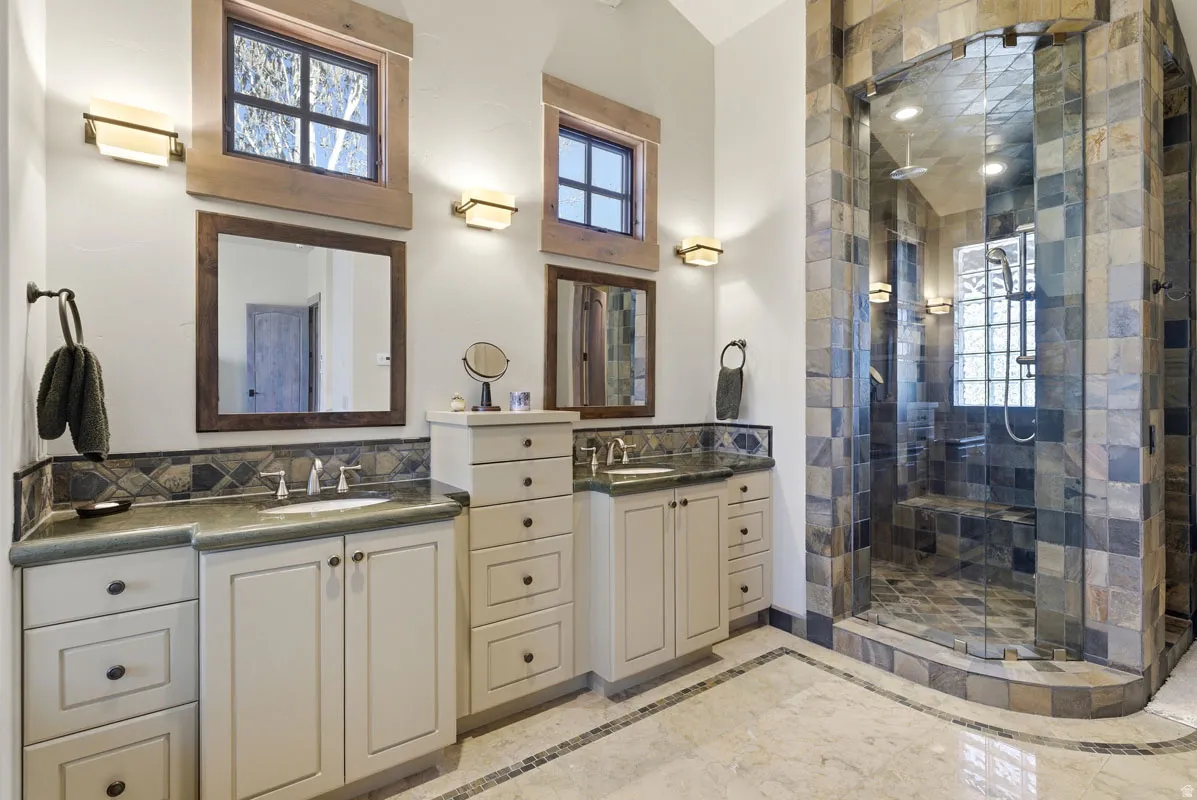 Spacious bathroom featuring a glass-enclosed shower with stone tile surround, dual vanity areas with undermount sinks, and light-toned cabinetry