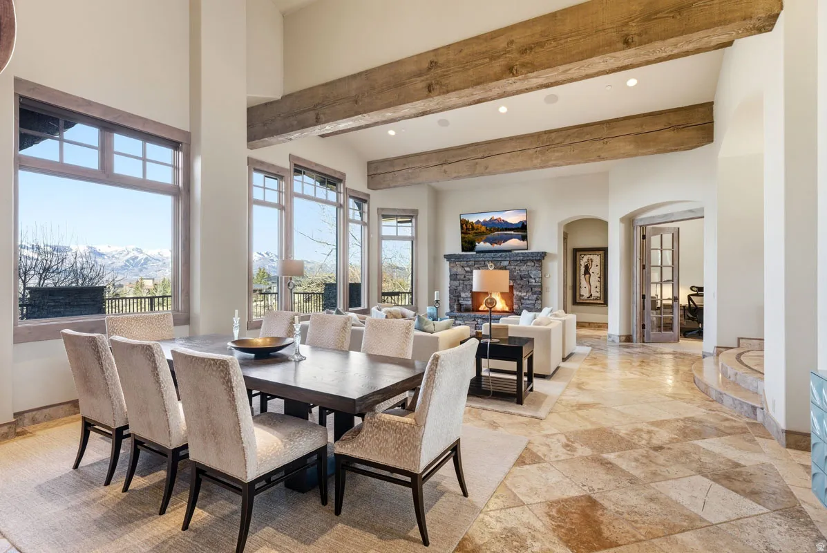 Open-concept living space featuring high ceilings with exposed wood beams, a stone fireplace, and expansive windows showcasing a mountain backdrop