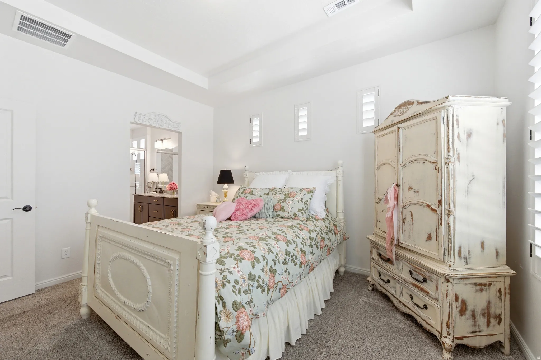 Bedroom featuring a tray ceiling, plush carpet flooring, white walls, and a doorway opening to an adjacent bathroom with a vanity and lighting