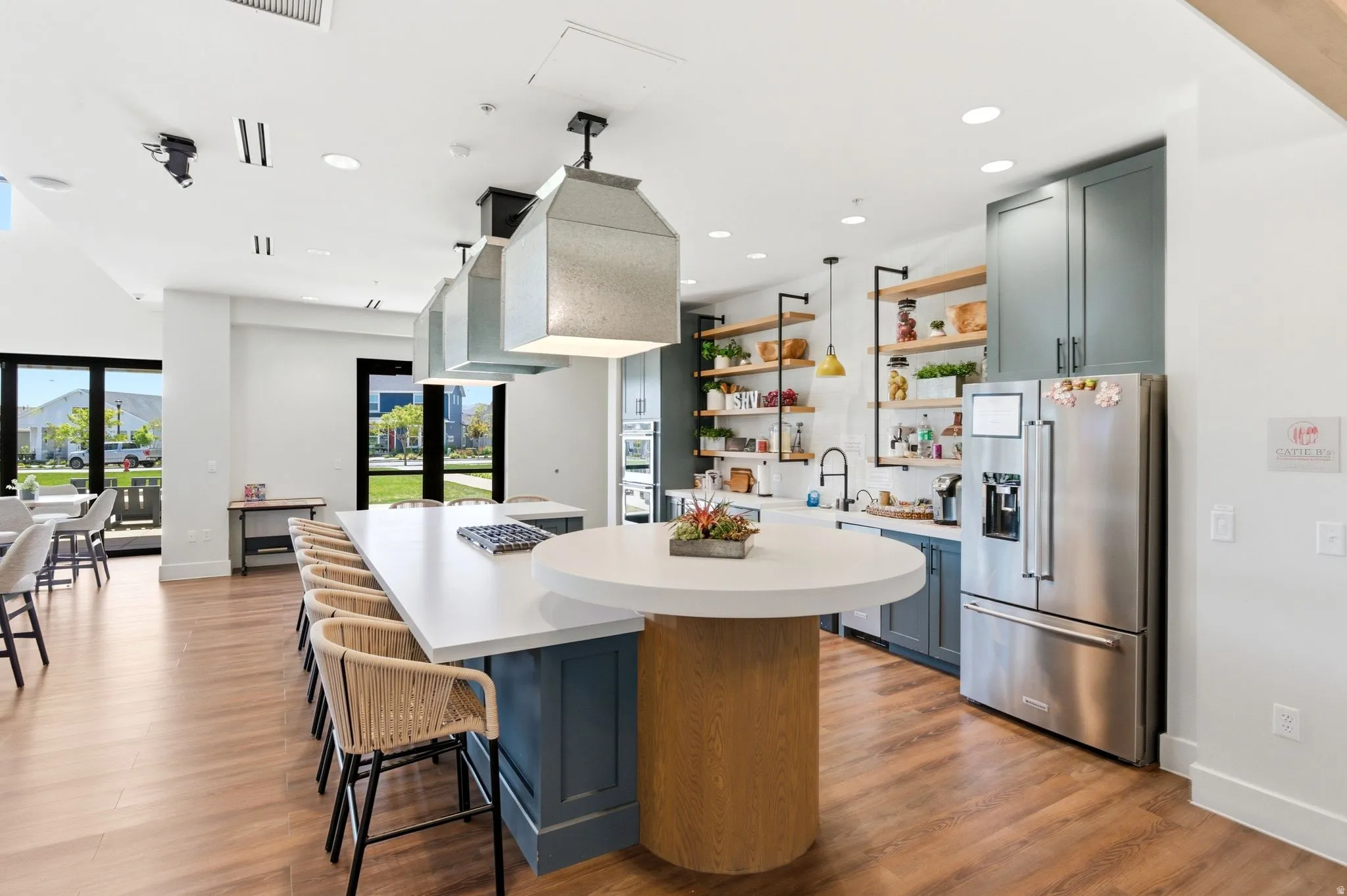 Expansive kitchen and dining area featuring a large central island with seating, warm wood-finish flooring, full-height windows, stainless steel appliances, and open shelving