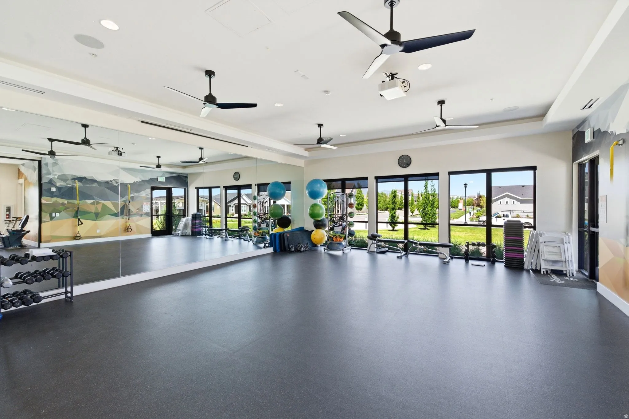 Spacious fitness room featuring expansive wall mirrors, durable black flooring, multiple ceiling fans, and large windows