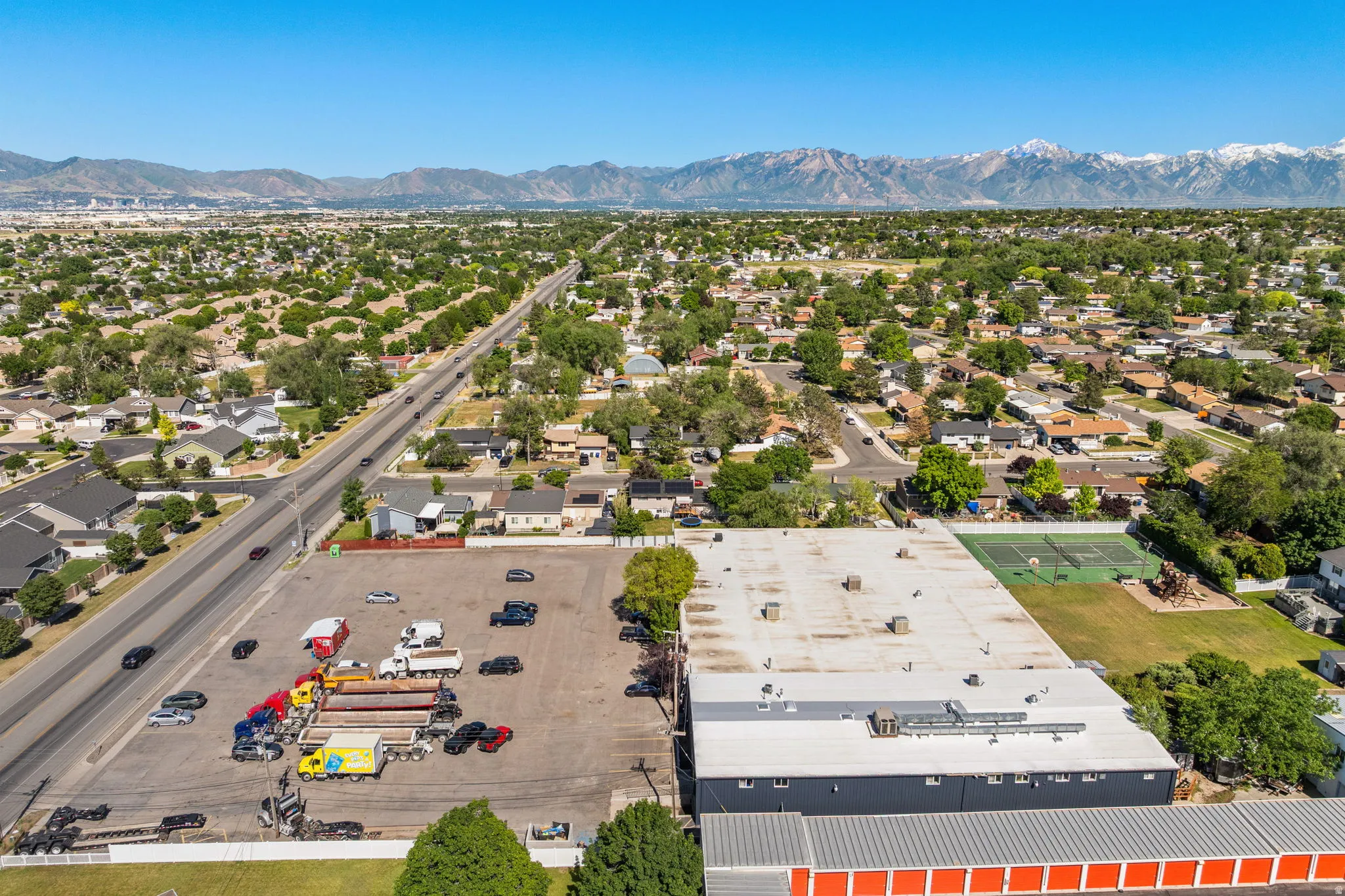 Commercial property featuring a large asphalt parking lot, an expansive industrial building with a flat roof, and an additional structure with orange-fronted storage units