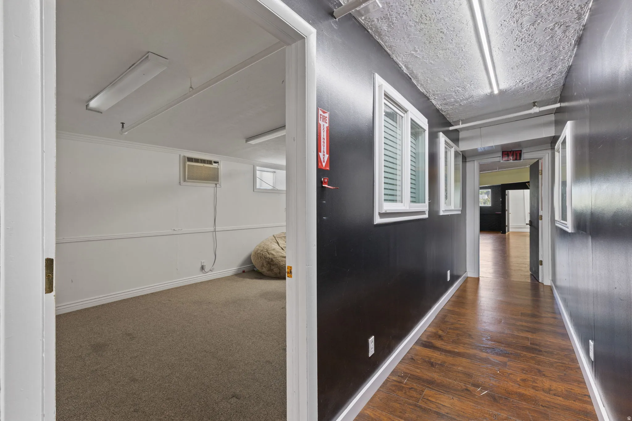 Hallway featuring wood-finish flooring and multiple windows, with a separate room featuring carpeted flooring and an air conditioning unit