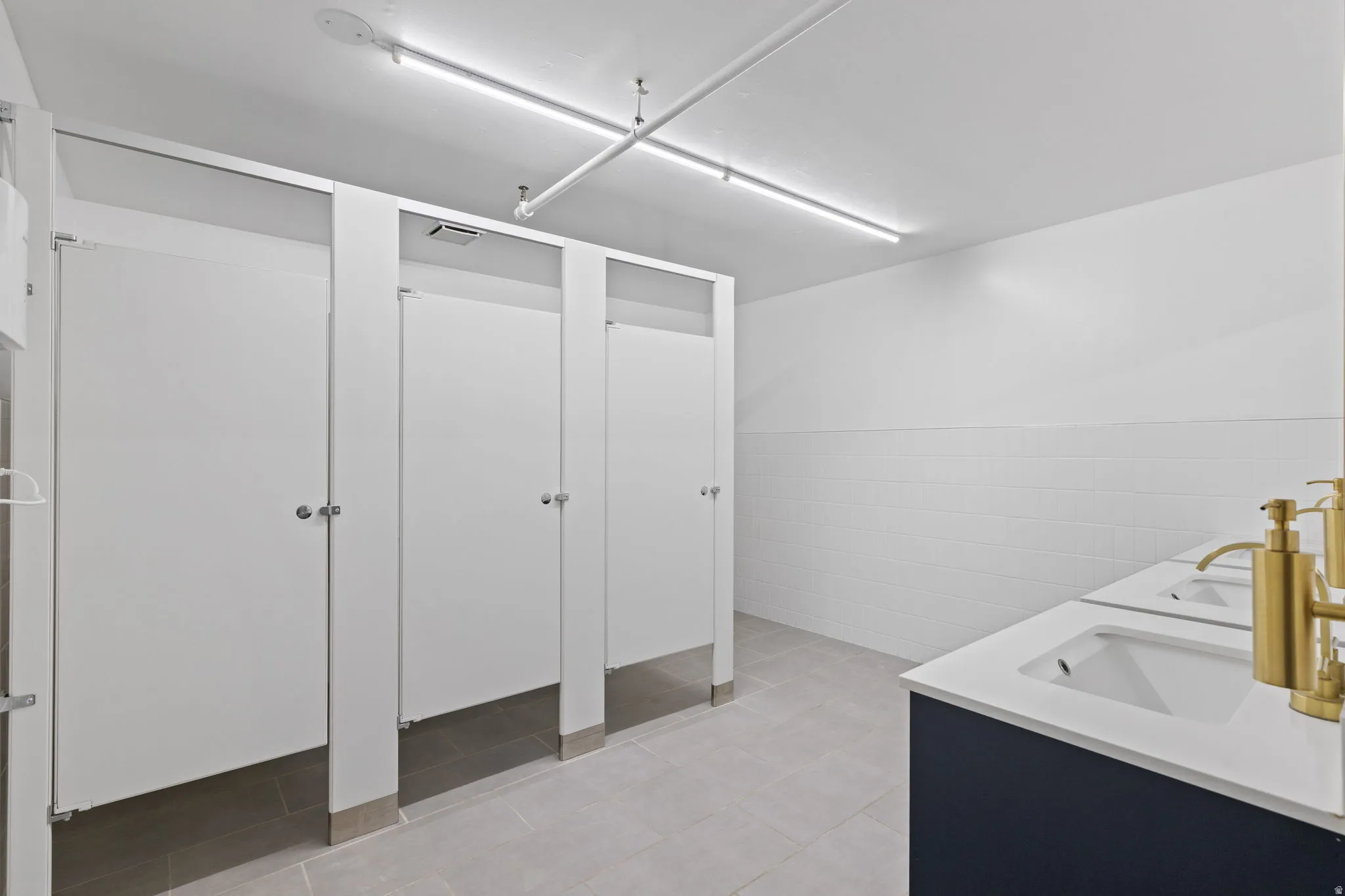 Commercial restroom with multiple stalls featuring white paneling, gray floor tiles, and white subway tile wainscoting