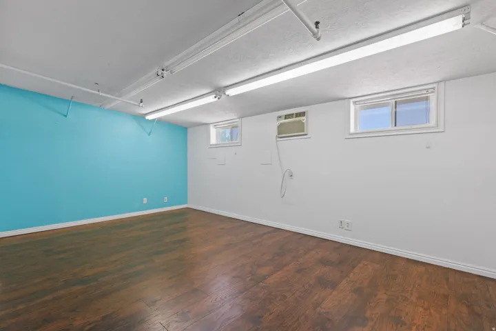 Spacious room featuring wood-finish flooring, a vibrant accent wall, recessed windows, an exposed ceiling with fluorescent lighting, and a wall-mounted air conditioning unit