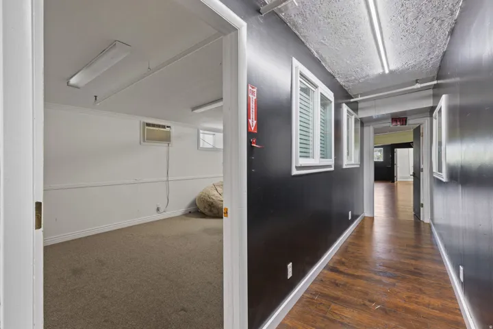 Hallway featuring wood-finish flooring and multiple windows, with a separate room featuring carpeted flooring and an air conditioning unit