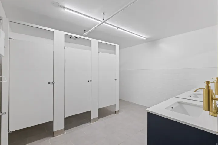 Commercial restroom with multiple stalls featuring white paneling, gray floor tiles, and white subway tile wainscoting