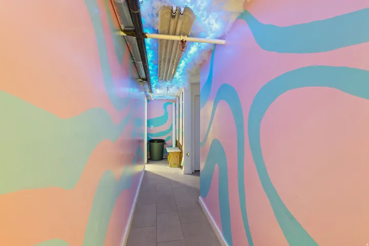 Hallway featuring light-toned tile flooring, brightly painted walls with abstract patterns, exposed ceiling pipes, and cloud-like ceiling decor with accent lighting