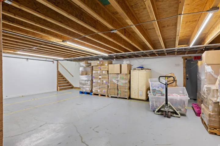 Spacious unfinished basement offering concrete flooring, exposed wood joists, fluorescent lighting, a visible staircase, and an exit door