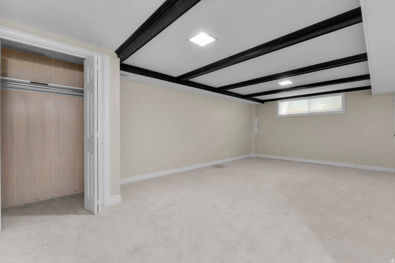 Spacious room featuring light-colored carpeting, neutral painted walls, and a distinctive ceiling with dark exposed beams and recessed lighting