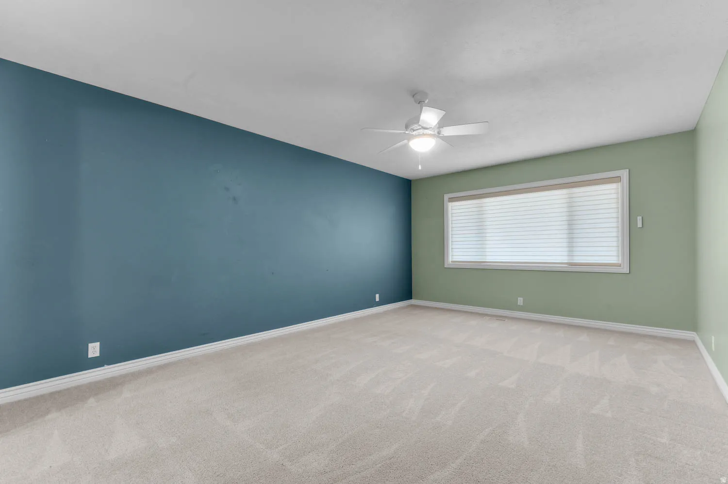Spacious room featuring neutral carpeting, a large window with blinds, a ceiling fan with integrated lighting, and walls in contrasting blue and green tones