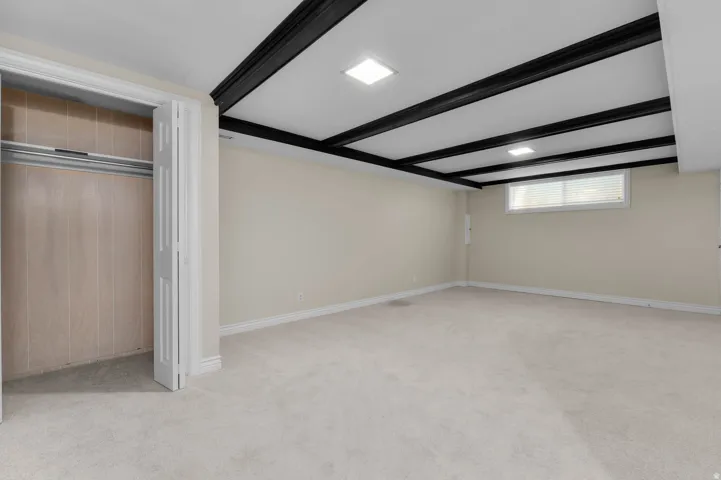 Spacious room featuring light-colored carpeting, neutral painted walls, and a distinctive ceiling with dark exposed beams and recessed lighting