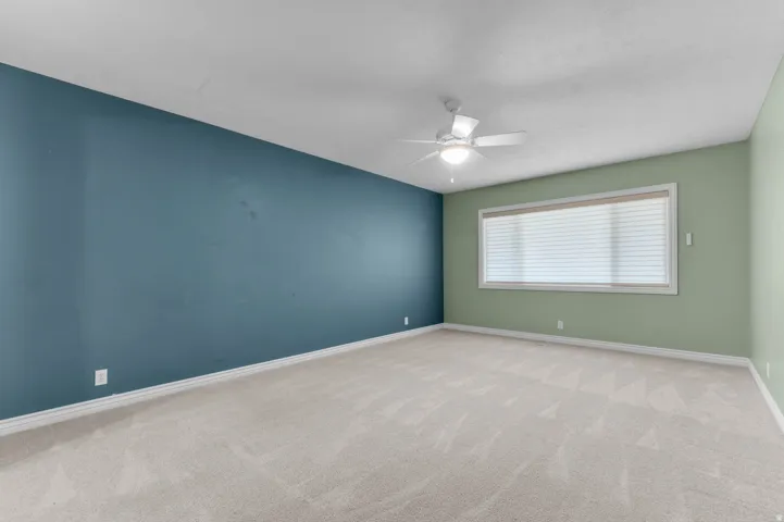 Spacious room featuring neutral carpeting, a large window with blinds, a ceiling fan with integrated lighting, and walls in contrasting blue and green tones