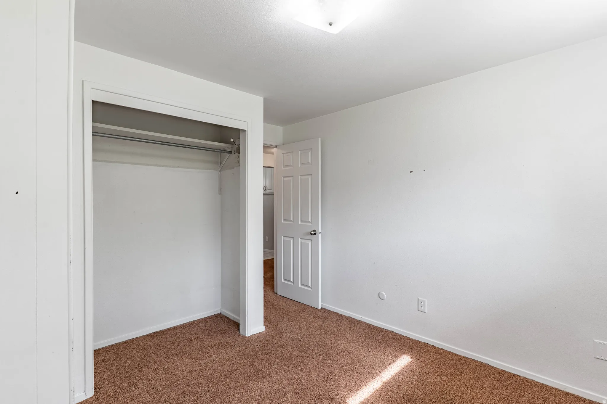Neutral white walls complement the brown carpet flooring in this room featuring an open closet with a hanging rod