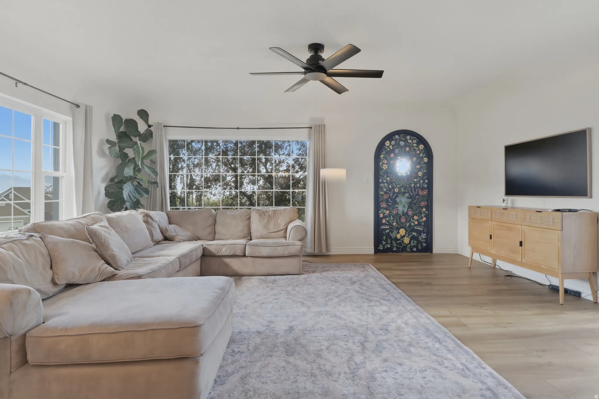 Spacious living area featuring light wood-finish flooring, a contemporary ceiling fan, and large windows providing ample natural light