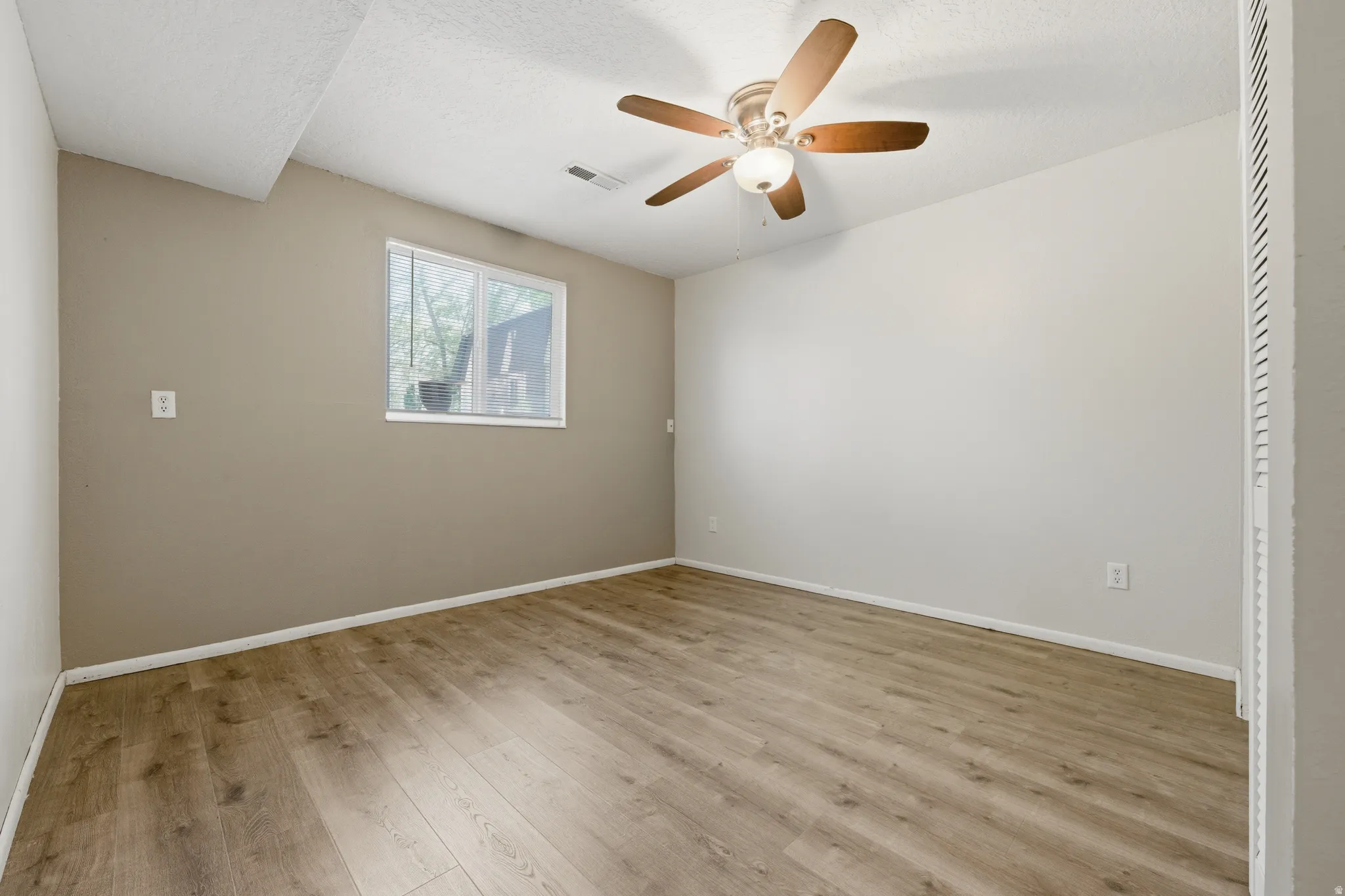 Spacious room featuring wood-finish flooring, a ceiling fan with integrated lighting, and a window for natural illumination