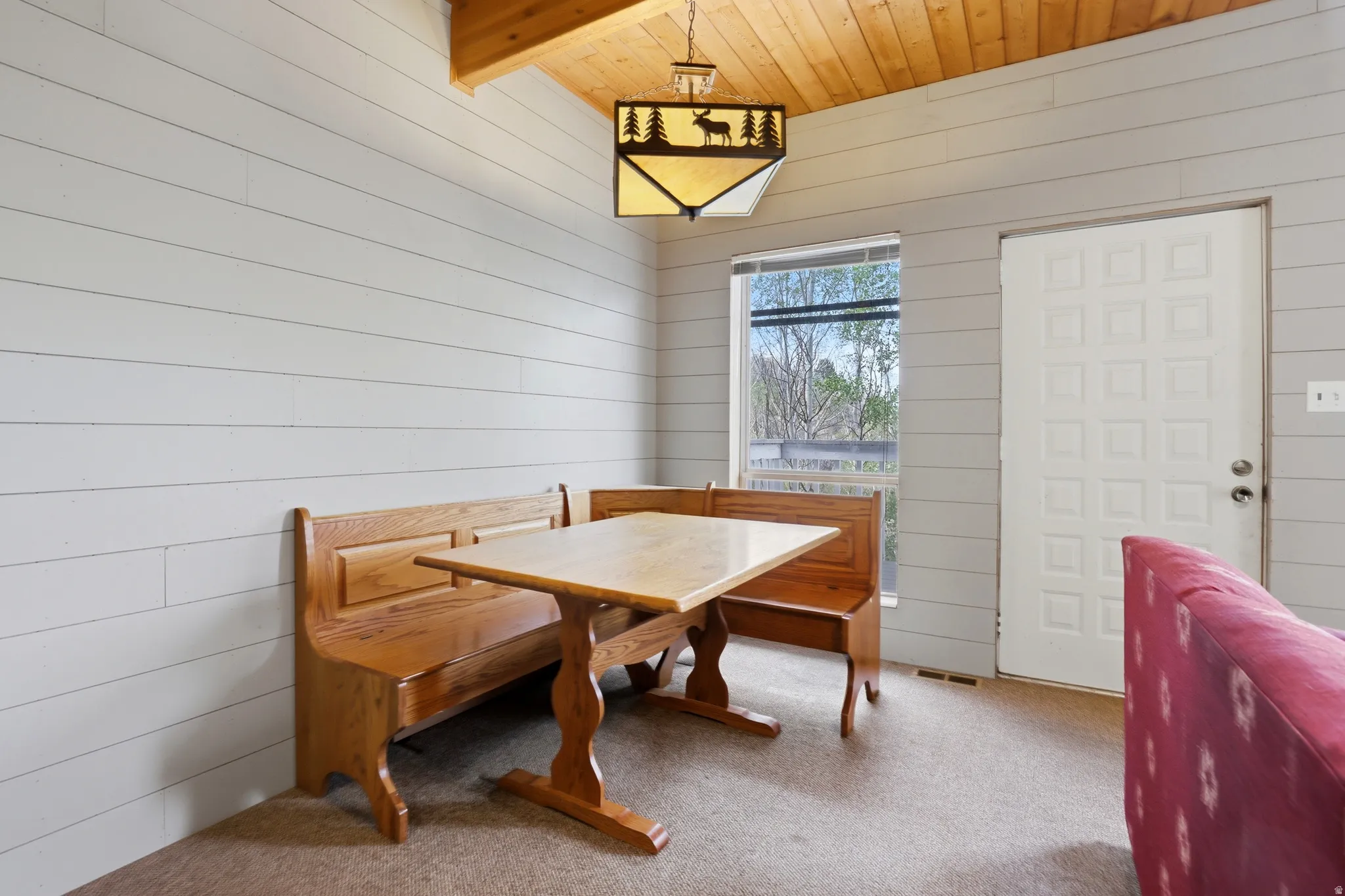 Vaulted wood-paneled ceiling with a decorative pendant light fixture, shiplap-style walls, carpeted flooring, and a window providing natural illumination