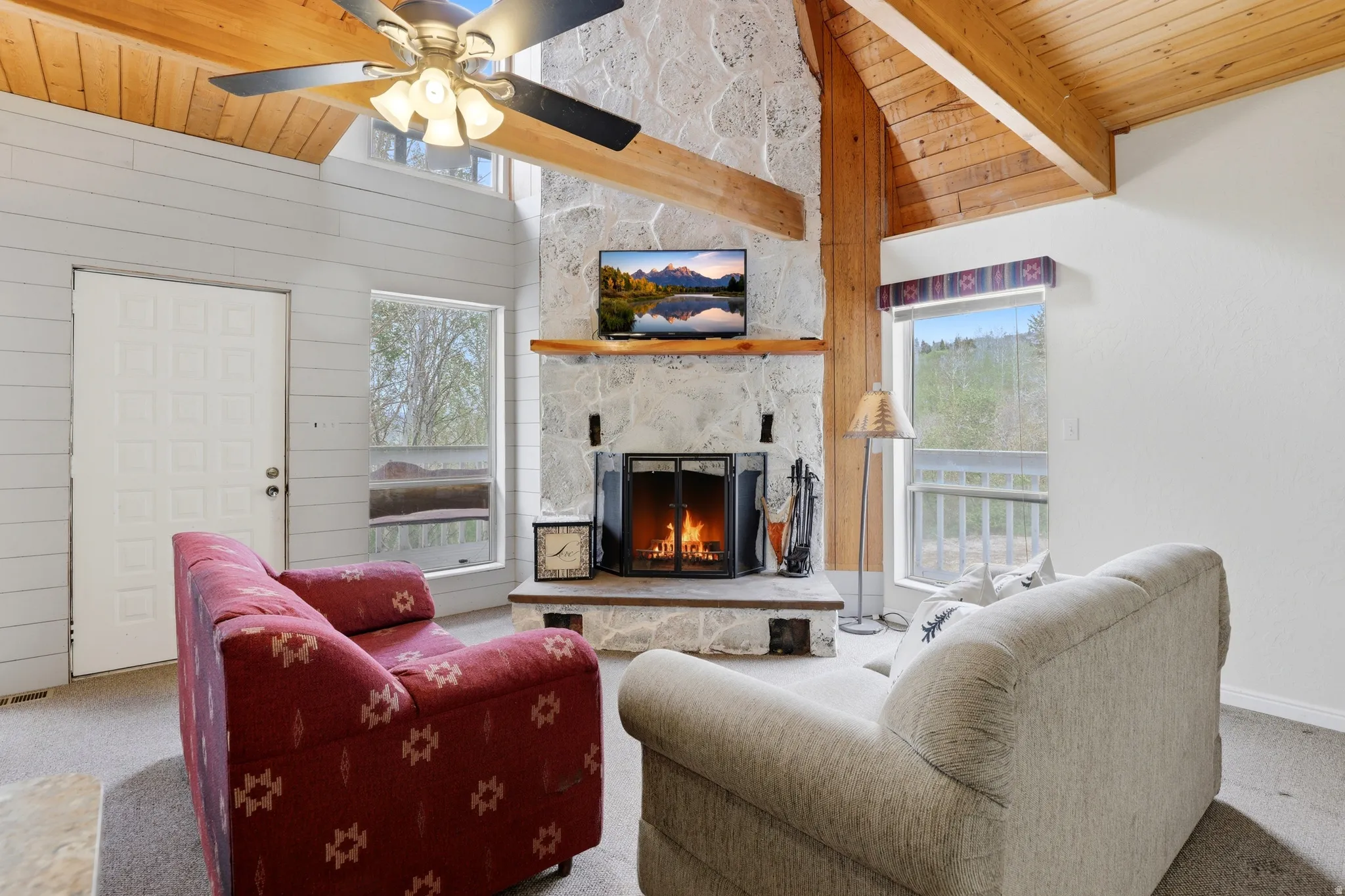 Vaulted wood-paneled ceiling with exposed beams, stone fireplace with wood mantel, white paneled walls, and multiple windows