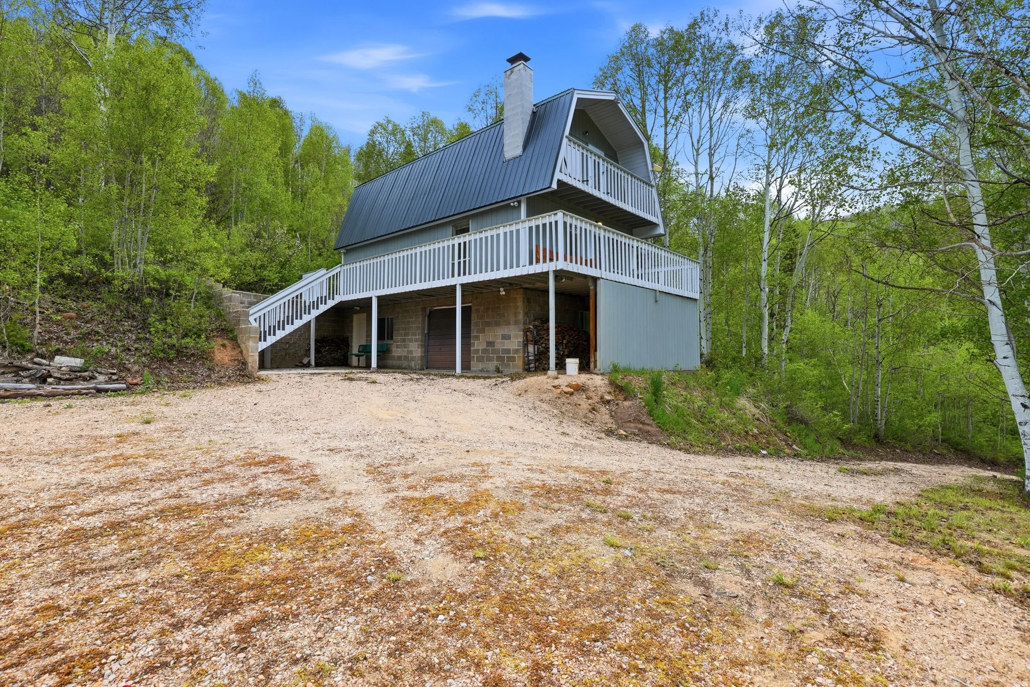Gambrel-style residence featuring a metal roof, an upper-level balcony, a wraparound deck with white railings, a block foundation, and surrounding mature trees