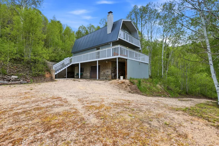 Gambrel-style residence featuring a metal roof, an upper-level balcony, a wraparound deck with white railings, a block foundation, and surrounding mature trees