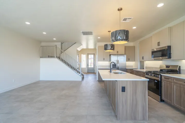 Open-concept living space with light-tone tile flooring, featuring a modern kitchen island with integrated sink, stainless steel appliances, and a staircase with light wood banisters