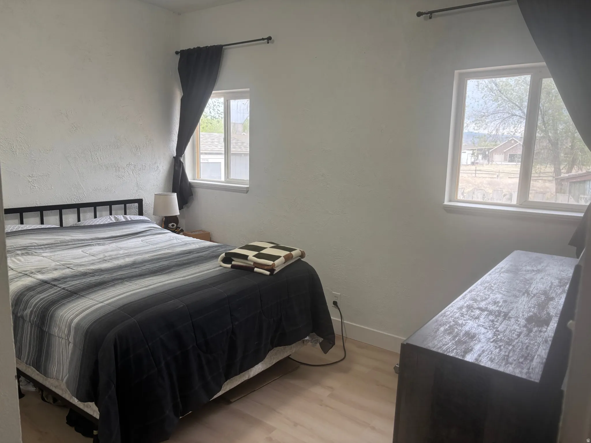 Bright bedroom featuring light wood-finish flooring, white textured walls, and two windows with white frames