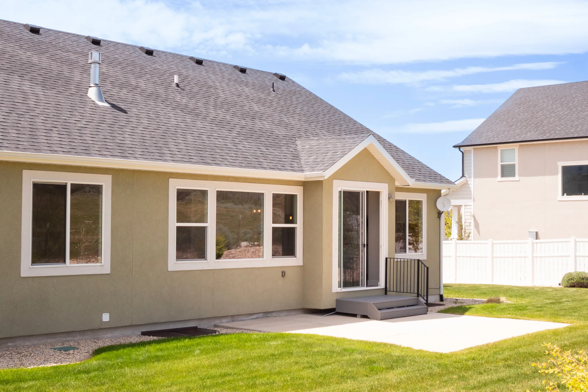 Rear exterior featuring a large concrete patio, sliding glass door with a small landing and metal railing, multiple windows with white frames, and a shingle roof