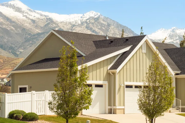 Residence with stucco and vertical siding, featuring a dark shingle roof, white garage doors, and a snow-capped mountain backdrop