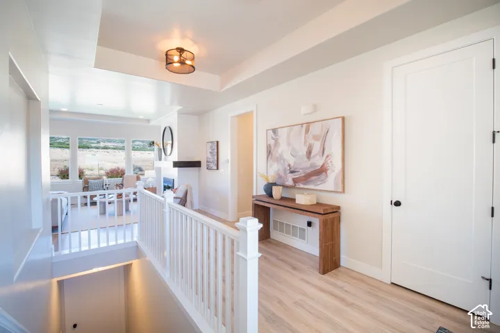 Light wood-finish flooring, white wood railing, tray ceiling with a modern light fixture, and a built-in fireplace with mantle