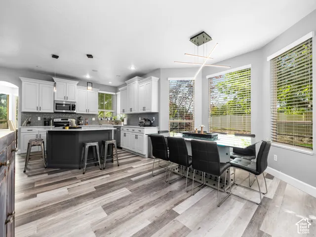Contemporary kitchen and dining area featuring light-toned wood-finish flooring, a dark kitchen island, white cabinetry, and a textured tile backsplash